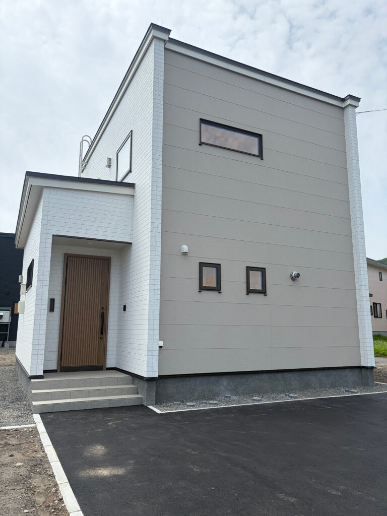 白いタイル調とグレージュの外壁、木目の玄関ドアを組み合わせた、登別市新生町モデルハウスのナチュラルな外観